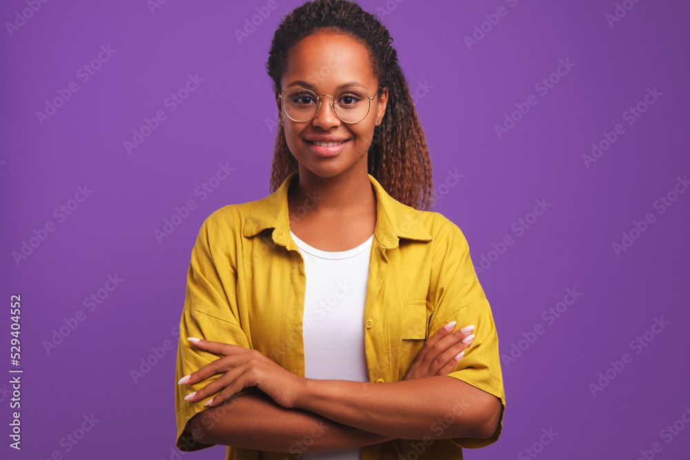 Young successful positive African American woman student arms crossed in front of chest looks at camera demonstrating purposefulness and self-confidence stands on studio plain background Young successful positive African American woman student arms crossed in front of chest looks at camera demonstrating purposefulness and self-confidence stands on studio plain background