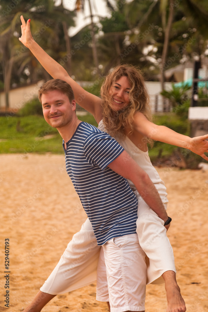 Happy lovely couple having fun on tropical sandy beach at palm trees background. Man giving piggyback ride to his woman and laughing. Joyful couple enjoying summer at ocean coastline. Copy text space