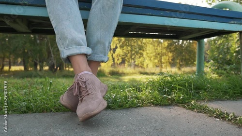 Girl dangling her feet in jeans and pink shoes. Woman swinging legs in boredom, sitting at park on a bench.