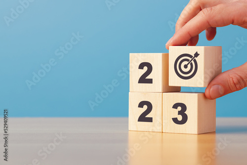 New year 2023. Hand putting wooden block with target icon on stack of wooden blocks with numbers 2023 on blue background copy space.