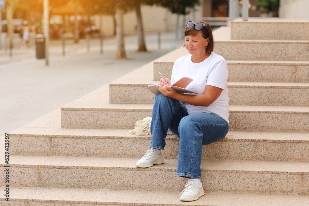 Obraz premium mature woman works remotely, higher education for adults, casually female writes and makes note in a notebook sitting on stairs outdoors, freelancer. College study program, academic educational course