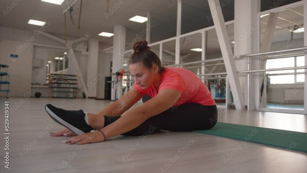 A beautiful young girl stretches in the gym and does incline to the leg while sitting on the mat. Slow motion A beautiful young girl stretches in the gym and does incline to the leg while sitting on the mat. Slow motion