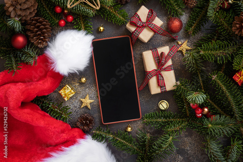 Mobile phone between Christmas decorations, Santa Claus hat, gift boxes and green fir branches