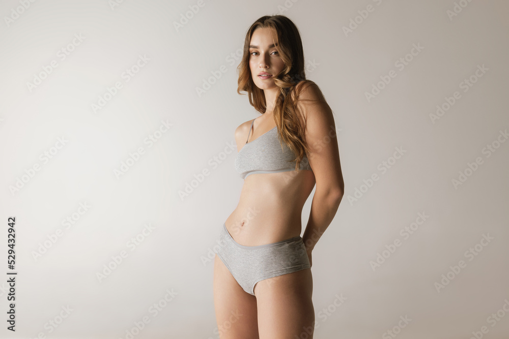 Obraz premium Studio shot of young charming slim girl in grey cotton underwear isolated over gray background. Wellness, fitness, diet, fashion, natural beauty of female body