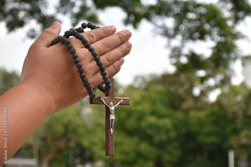 Wooden cross and wooden rosary are held in hands of young asian Catholic prayer while praying in the temple park area. Wooden cross and wooden rosary are held in hands of young asian Catholic prayer while praying in the temple park area.