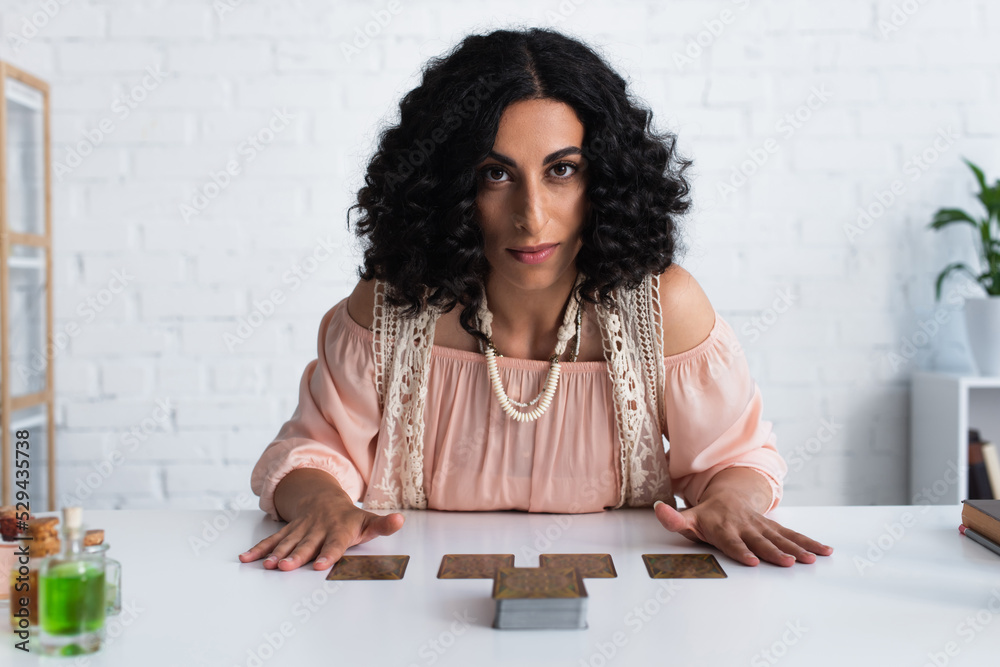 KYIV, UKRAINE - JUNE 29, 2022: young predictor looking at camera while sitting near tarot cards. KYIV, UKRAINE - JUNE 29, 2022: young predictor looking at camera while sitting near tarot cards.