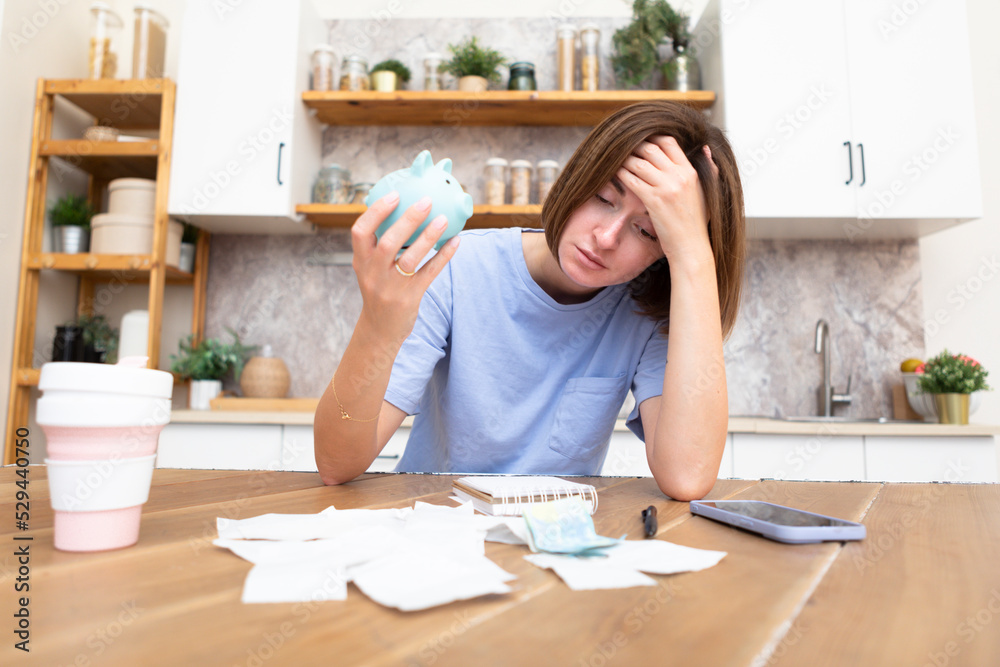Unhappy and depressed woman sitting in the kitchen at the table, holding piggy bank and looking at a lot of bills and checks. Taxes and debts, family budget and spending concept Unhappy and depressed woman sitting in the kitchen at the table, holding piggy bank and looking at a lot of bills and checks. Taxes and debts, family budget and spending concept