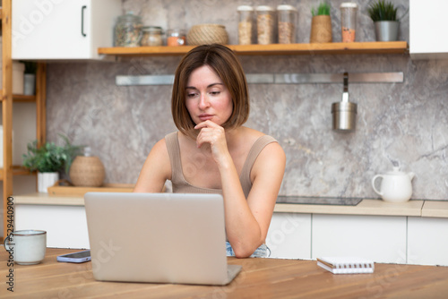 Young attractive woman sitting at the table and working on laptop in the kitchen at home 