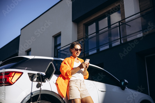 Woman charging electric car and using phonr by her house photo