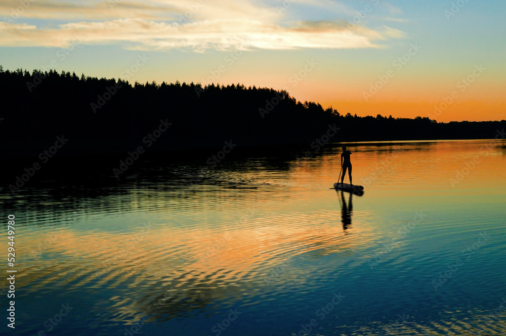 Silhouette of a girl on a SUP board on the river. Evening, beautiful sunset of pink, yellow and blue hues, twilight, picturesque landscape. Postcard, wallpaper, background. Summer, water sports.