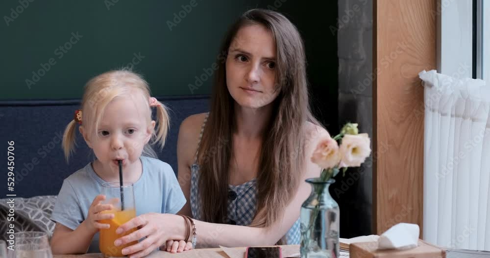 Young mother and little blonde daughter relax in cafe. Girl drinks cocktail or limonade through straw. Family rest
