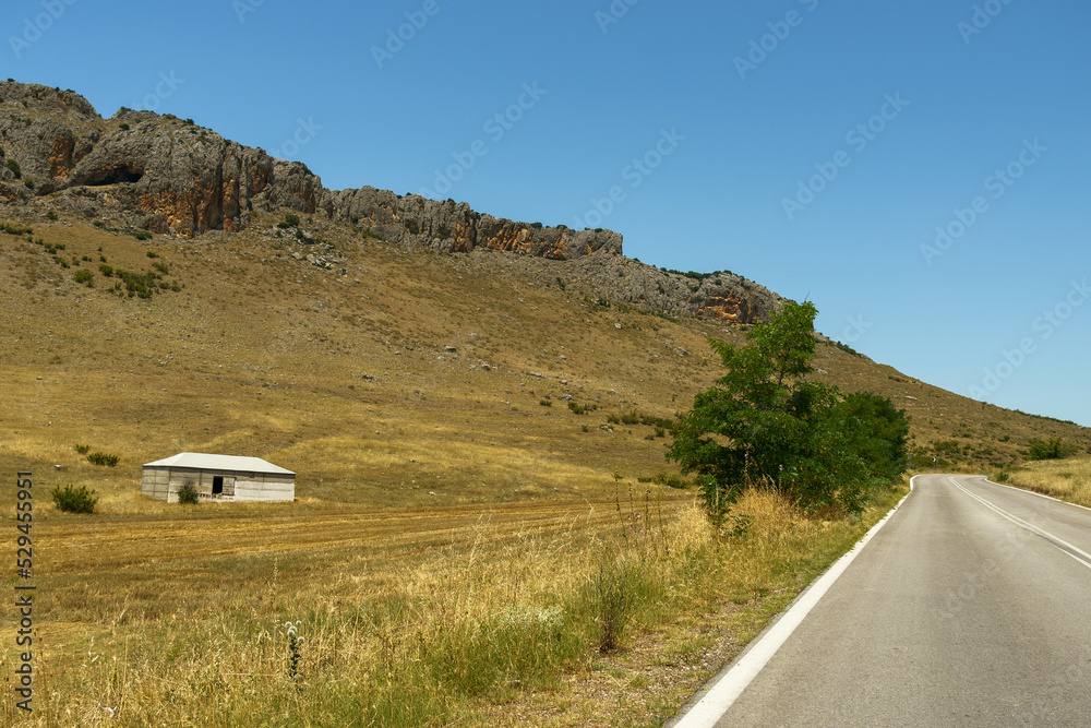Obraz premium Asphalt road leading through rocky mountains and an abandoned house.