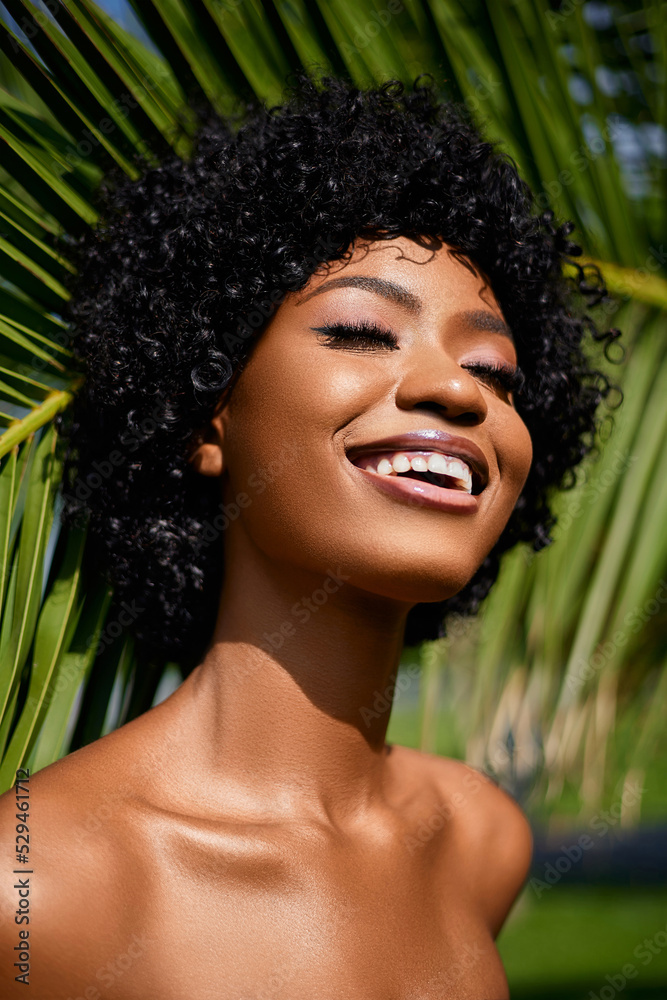 Overjoyed African American model with a curly hair and bare shoulders smiling and posing against palms tree. Natural skin care concept Overjoyed African American model with a curly hair and bare shoulders smiling and posing against palms tree. Natural skin care concept