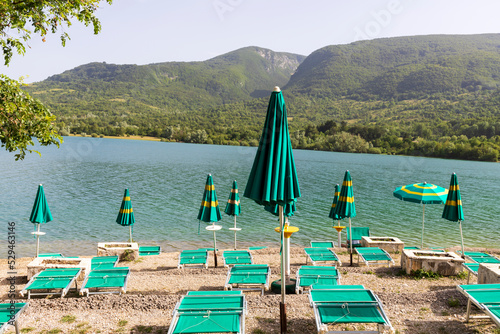 Fototapeta Naklejka Na Ścianę i Meble -  The beach with sunbeds and umbrellas on a lake and mountains in the background. Barrea lake, Abruzzo, Italy.