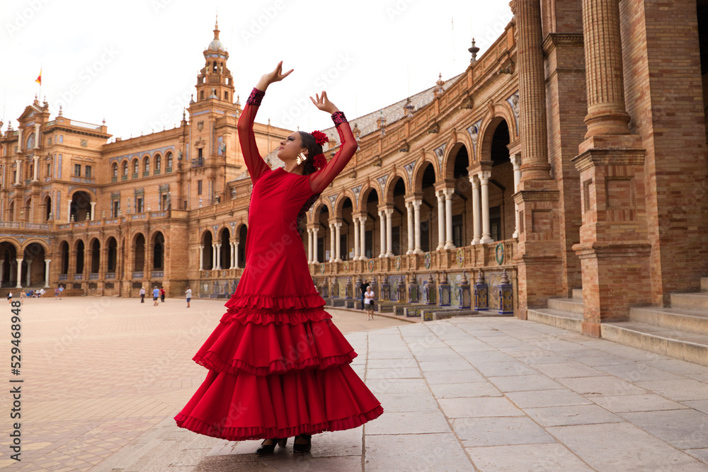 Naklejka premium Beautiful teenage woman dancing flamenco in a square in Seville, Spain. She wears a red dress with ruffles and dances flamenco with a lot of art. Flamenco cultural heritage of humanity.