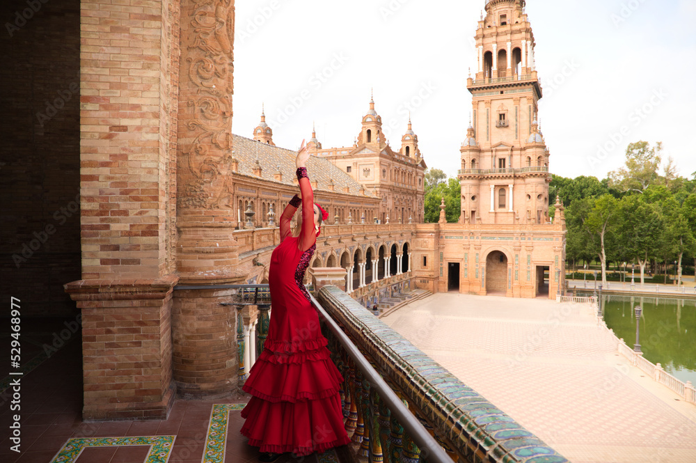 Fototapeta premium Beautiful teenage woman dancing flamenco in a square in Seville, Spain. She wears a red dress with ruffles and dances flamenco with a lot of art. Flamenco cultural heritage of humanity.