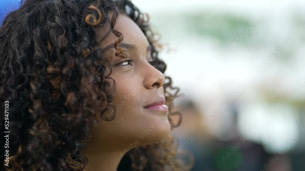 Happy young African American woman standing outdoors looks up at sky smiling. A grateful black 20s adult girls opening eyes