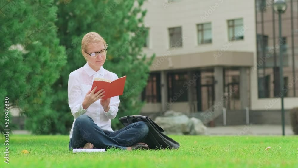 Middle aged blonde woman in glasses and white shirt flipping through the organizer sitting on green lawn by eco friendly office building. Businesswoman in formal clothes working outdoors. Middle aged blonde woman in glasses and white shirt flipping through the organizer sitting on green lawn by eco friendly office building. Businesswoman in formal clothes working outdoors.