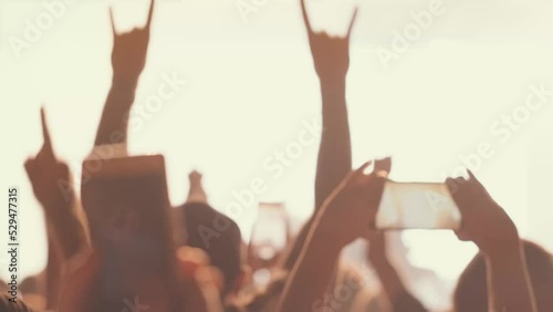 A blurry and illuminated dancing crowd. A crowd of fans standing near the stage applauding their favorite rock band. Concert in the city center. Young people with smartphones.