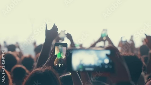 A blurry and illuminated dancing crowd. A crowd of fans standing near the stage applauding their favorite rock band. Concert in the city center. Young people with smartphones. photo