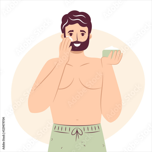 Skincare routine concept. Happy young man applying face moisturizing cream or lotion on cheek, handsome bearded male enjoying skincare procedure.