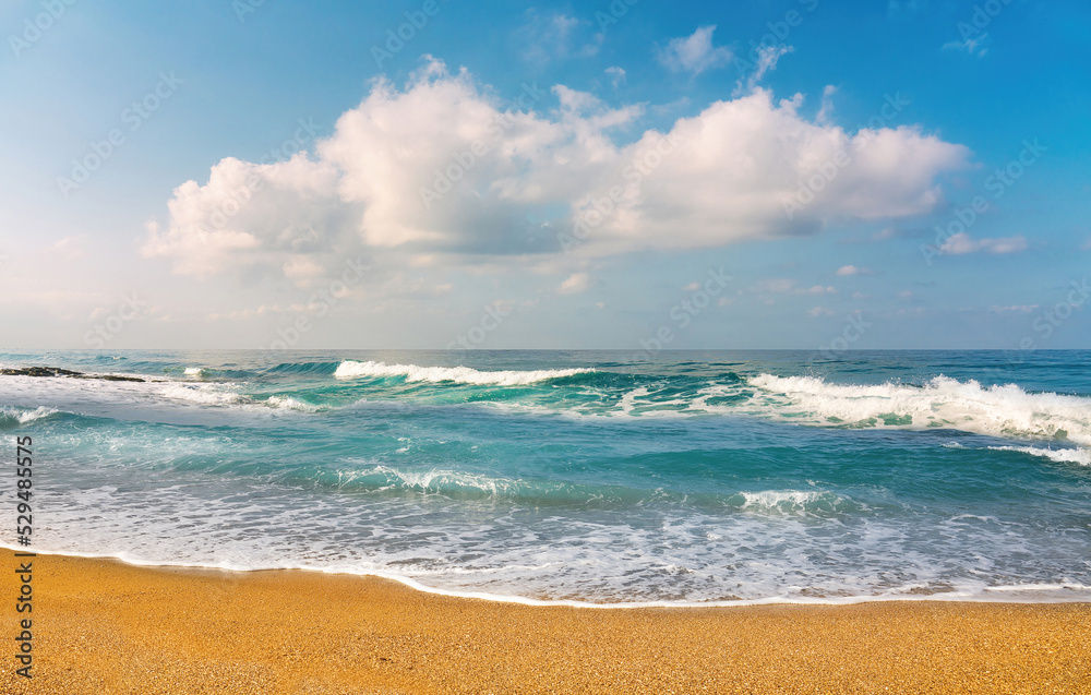 Beautiful natural seascape with turquoise sea, waves crashing on a sandy shore beach against a blue sky with clouds.