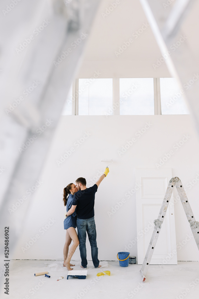 Young couple in love, the newlyweds are making repairs in an empty room, painting white walls, updating the house. A happy man and woman took out a mortgage and moved into a new house. Place for text 