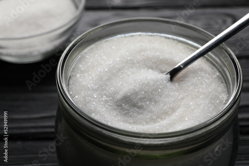 Granulated sugar and spoon in glass jar on black table, closeup