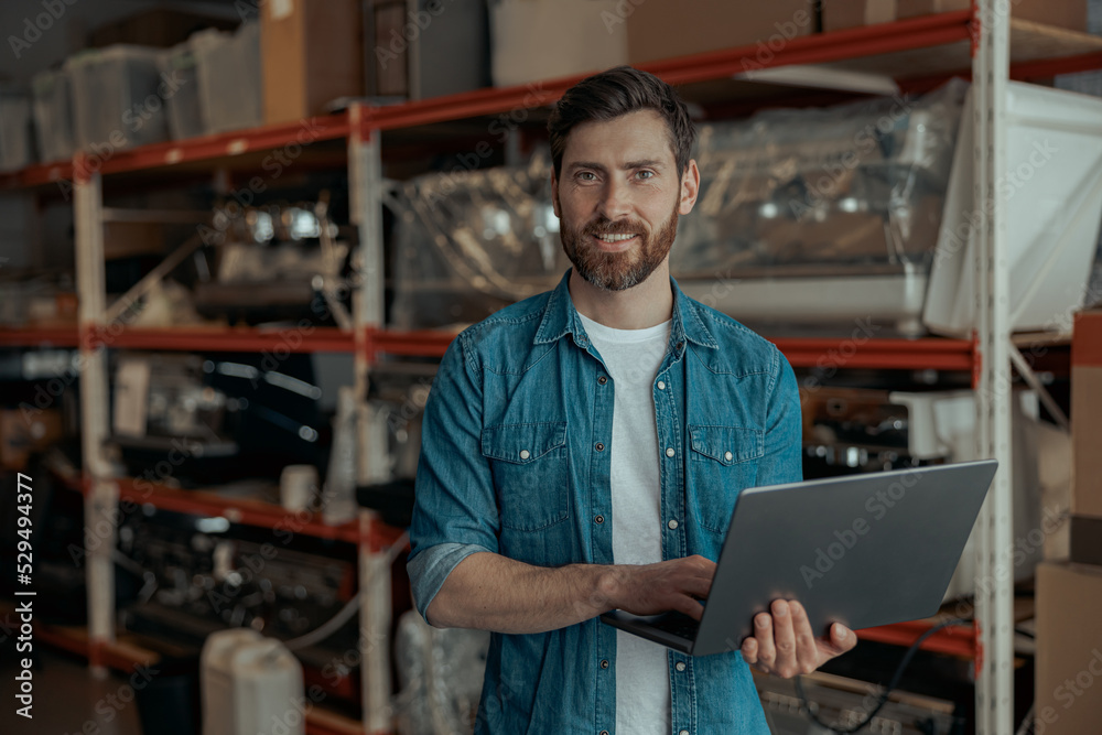 Male worker standing with laptop on warehouse with coffee machines Male worker standing with laptop on warehouse with coffee machines