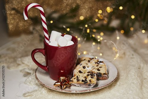 Aesthetic hot beverage red cup with marshmallows and Christmas stollen on ceramic plate - traditional holidays bread, candy cone stick. Festive Christmas background. Christmas tradition food. photo