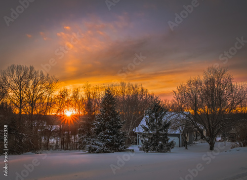 View of beautiful sunset with sunburst over Midwestern neighborhood in winter  snow on trees, buildings and the ground © Lana
