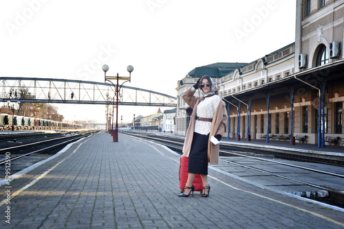 a young brunette with a scarf on her head in dark glasses and high heels is waiting for the arrival of a train on the railway