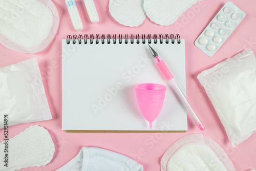 Top view photo of open planner over sanitary napkins, tampons, underwear and pills on isolated pastel pink background