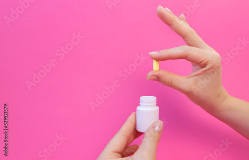 Photo of female hands holding a yellow pill and a white bottle of vitamins on a pink background