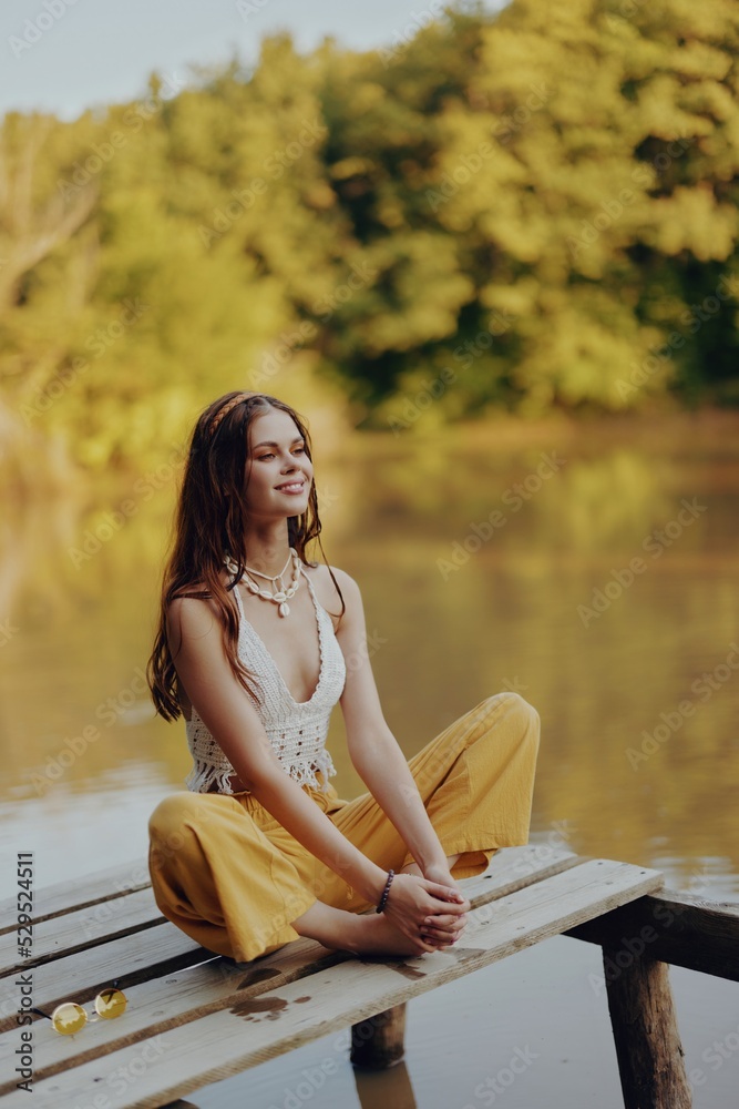A young happy woman with a hippie smile sits on the lake shore on a bridge and meditates in a lotus pose wearing eco clothing made of natural materials in harmony with nature in the fall