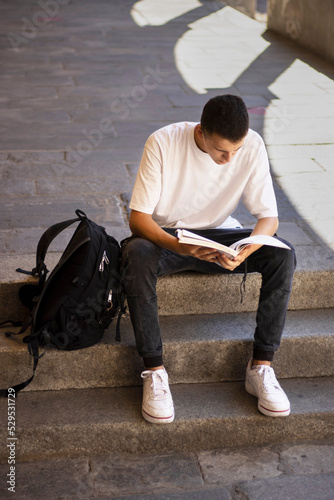 Young male student in a white t-shirt and dark jeans sitting on the stairs and reading a book , backpack, university, exams