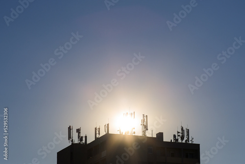 Many mobile transmitters on the roof of a high-rise building in the background with the sun
