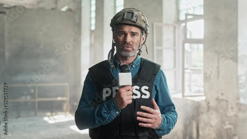 Close up portrait of middle-aged Caucasian handsome correspondent reporting live from place of battle. Man journalist in war zone talking in microphone looking at camera. War press photo