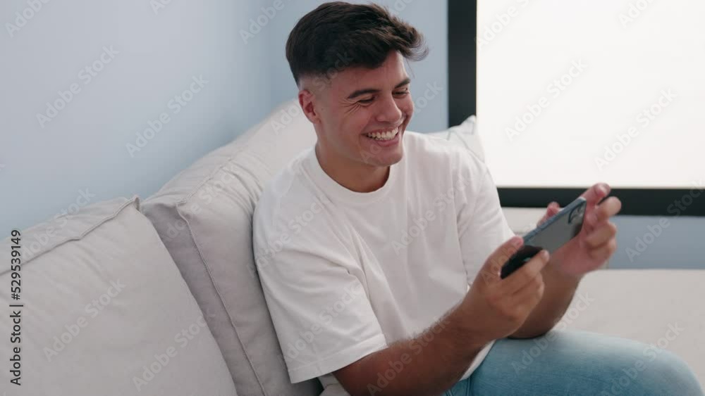 Young hispanic man watching video on smartphone sitting on sofa at home Young hispanic man watching video on smartphone sitting on sofa at home
