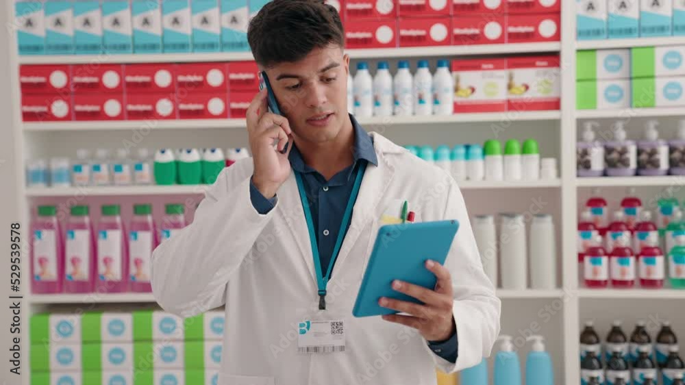 Young hispanic man pharmacist using touchpad talking on smartphone at pharmacy