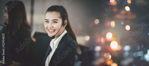 Asian woman in headset, sitting in office, working as operator of call center or support customer service, looking directly at the camera and smiling friendly. © May Chanikran