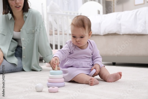 Cute baby girl playing with toy pyramid near mother on floor at home