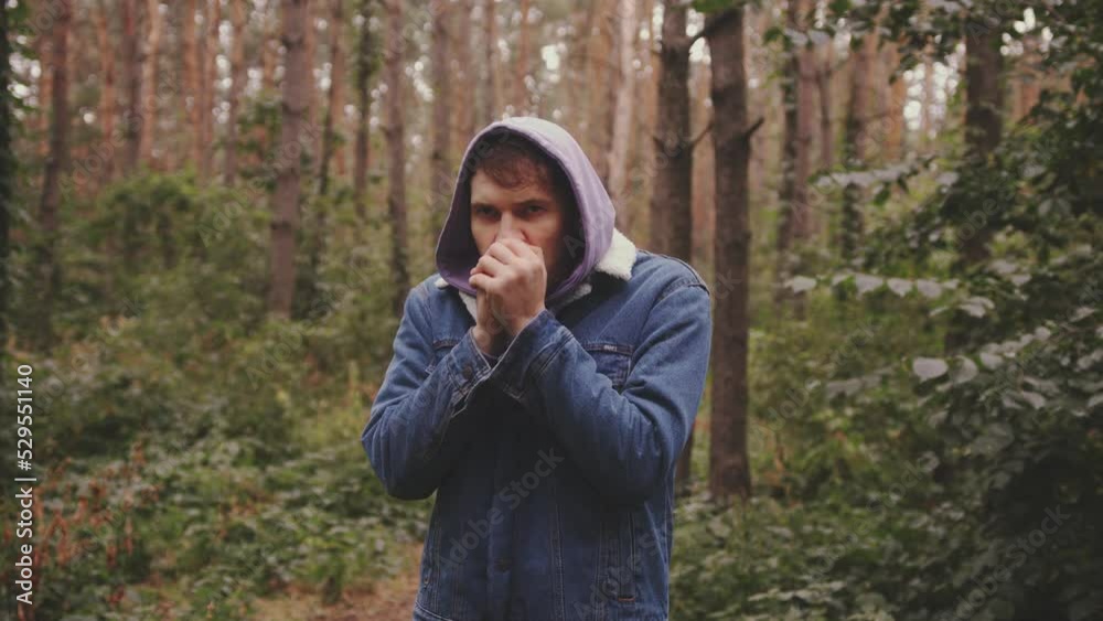Young frozen man clenching hands into fist, warming hands with his breath, standing in forest. Young frozen man clenching hands into fist, warming hands with his breath, standing in forest.