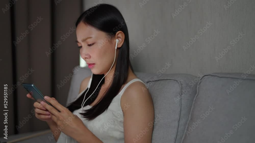 4K Depressed Asian woman sitting on couch by the window in living room listening sad song on earphones with crying. Loneliness girl living alone with sadness. Mental health and life problem concept.