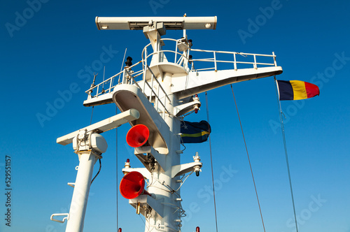 Fényképezés Ship's mast with flag of Antilles and Romania, navigation equipment, view from below against the sky