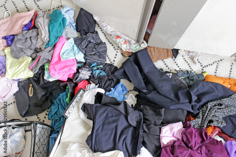 Above view of a messy bedroom floor with clothes overflowing from the wardrobe and storage boxes. Untidy cluttered woman's clothes scattered all over the floor.