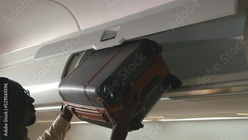 Passenger black man putting luggage on top shelf or cabin compartment on airplane, business man lift suitcases putting overhead locker on airplane, Travel concept photo