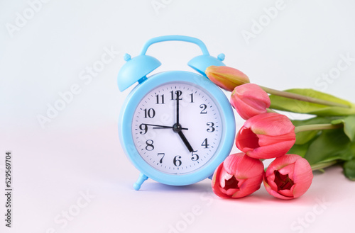 Blue alarm clock with spring flowers and space for text on light blue wooden background, flat lay.