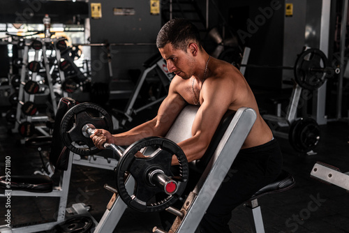 High quality photography. Latino man in profile with a barbell with weights doing curls to train the bicep. A muscular man doing exercises with a lot of effort in the gym.