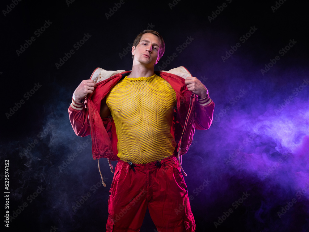 Obraz premium a young muscular man in a bright outfit poses in a pretentious pose, style and grotesque. Attractive guy with pumped-up arms in a mesh T-shirt and red pants, photo in smoke